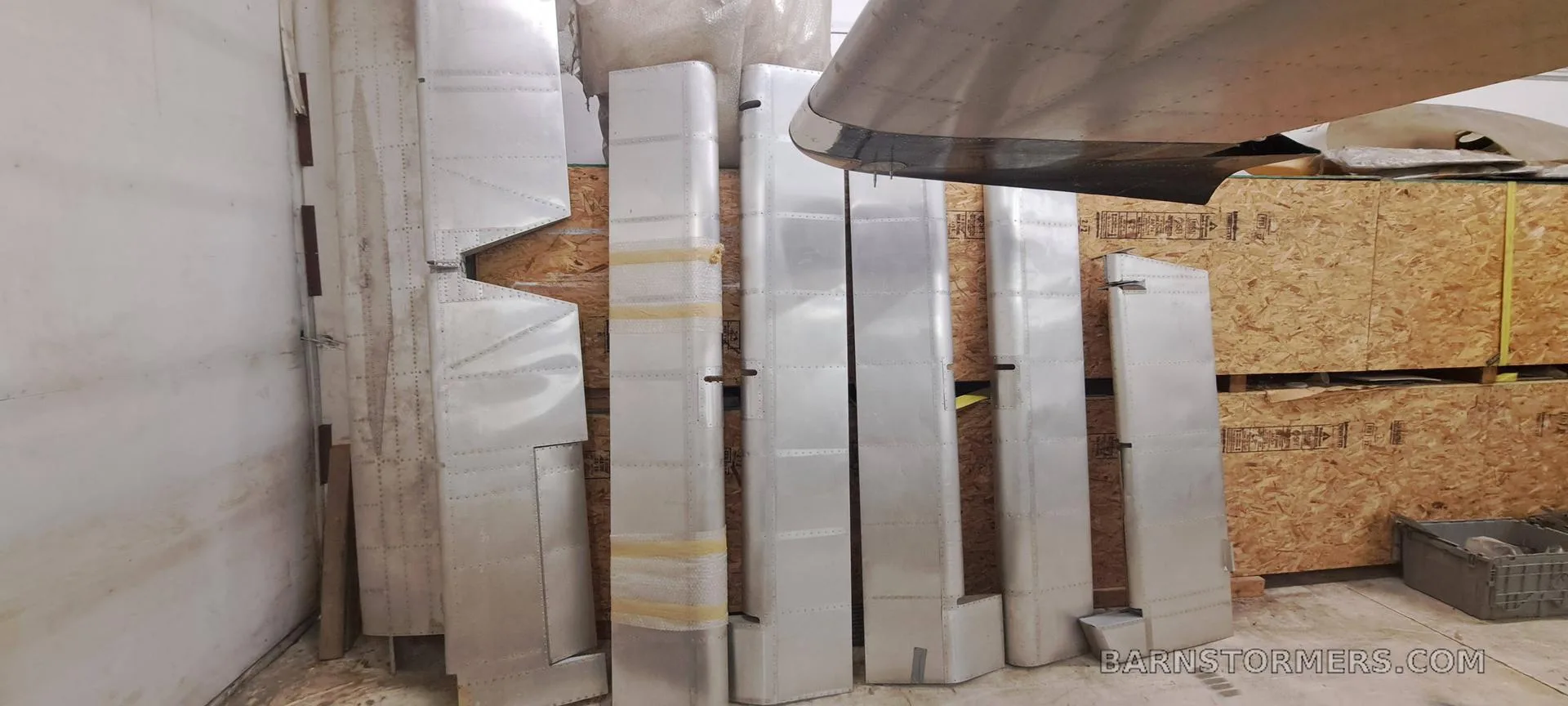 Various disassembled aircraft wing and tail sections made of aluminum, stored upright against a wall in a workshop.