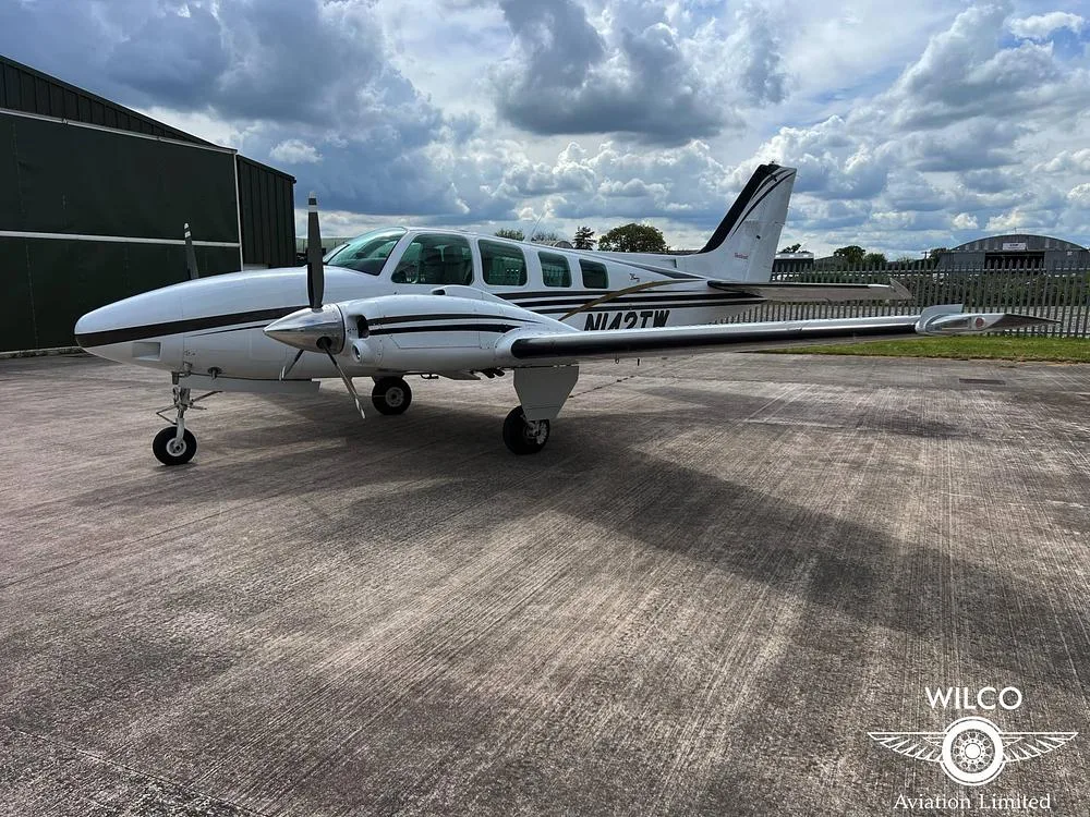 A white Beechcraft Baron N142TW with black and gold stripes parked on a tarmac under a cloudy sky.