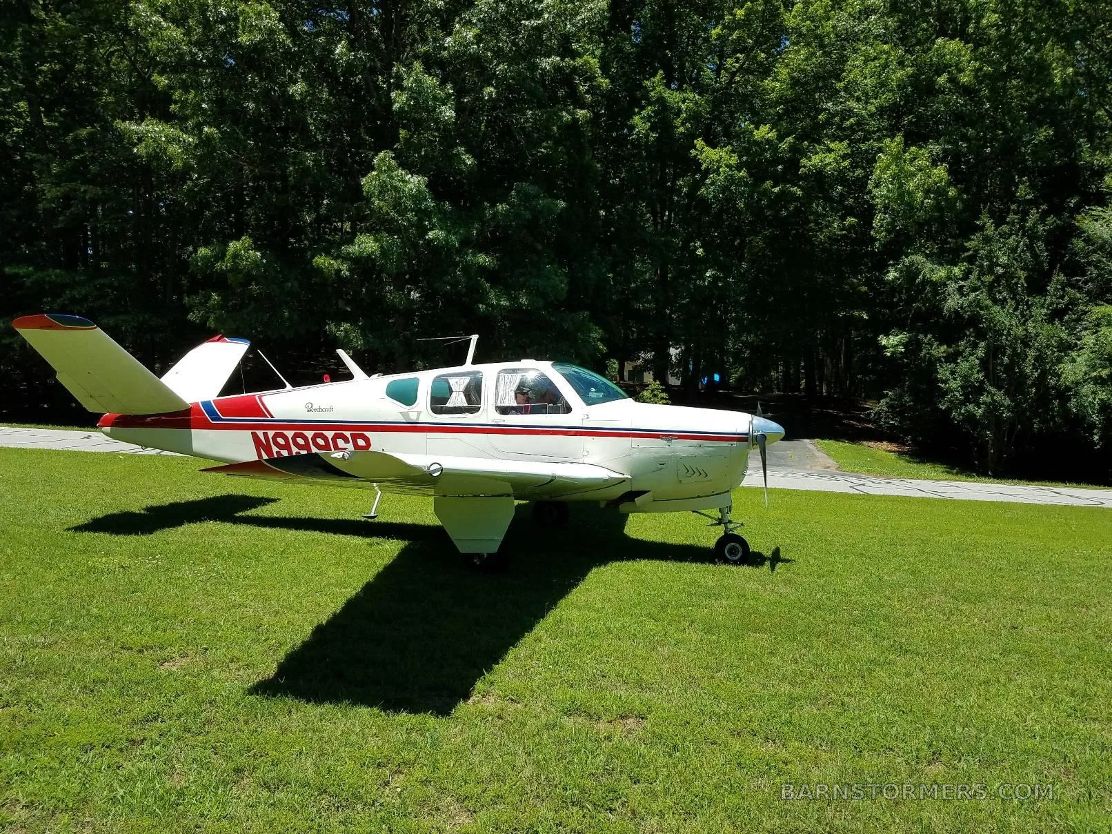 A white Beechcraft Bonanza with red and blue stripes, registration N999CR, parked on a grassy area with trees in the background.