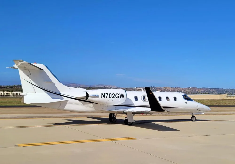 A white Learjet 35A aircraft with black stripes, bearing registration N702GW, parked on an airport tarmac under a clear blue sky.