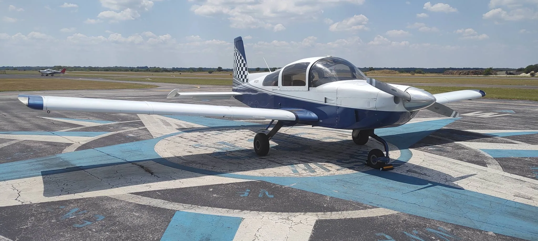 A blue and white single-engine aircraft, N74374, with a checkered tail, parked on an airfield compass rose.