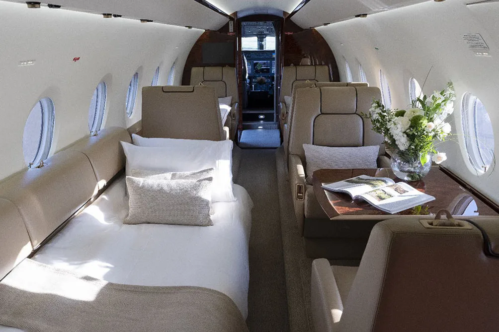 The interior cabin of a private jet configured with a bed, a dining table with flowers, and beige seating.