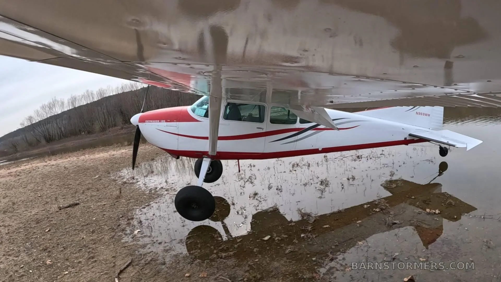 A low-angle view of a white and red Cessna 180 aircraft with tundra tires, reflecting in a large puddle on muddy ground.