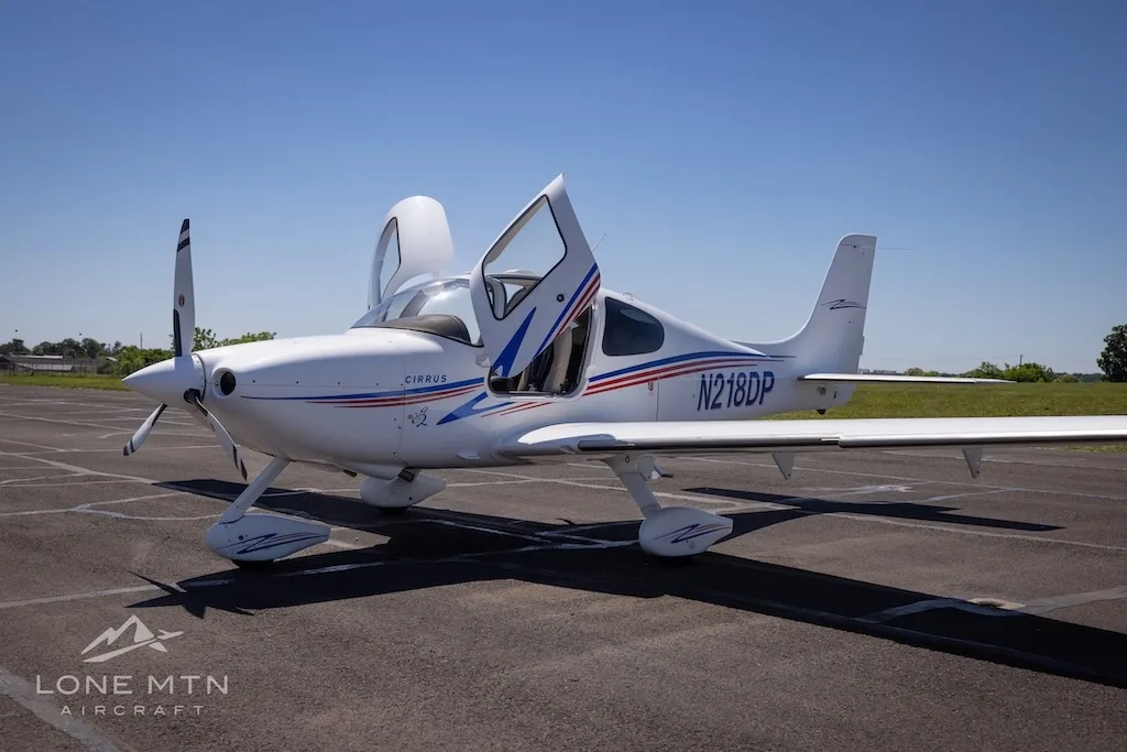 White Cirrus aircraft (N218DP) with blue and red stripes, doors open, parked on an asphalt runway under a clear sky.