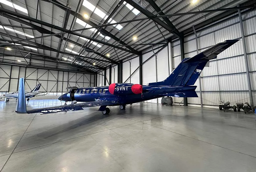 A dark blue private jet with registration G-SVNT and red engine covers parked in a hangar, viewed from the rear-right, with another jet in the background.