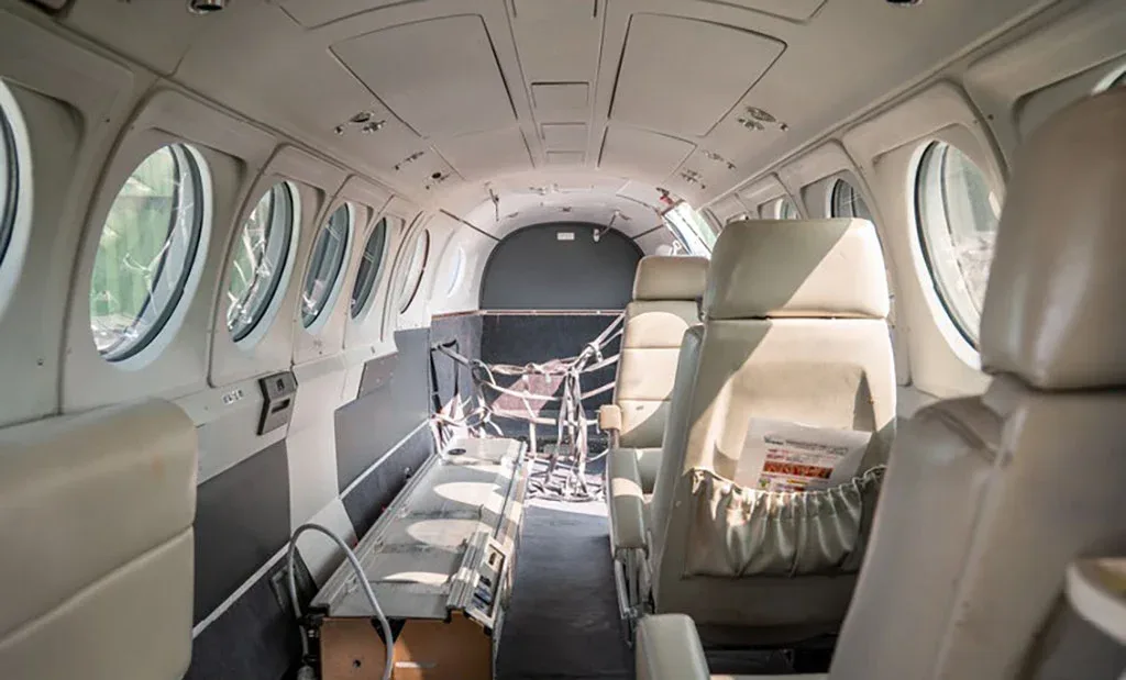 The interior cabin of a private aircraft with beige seats, large windows, and a view towards the rear cargo area.