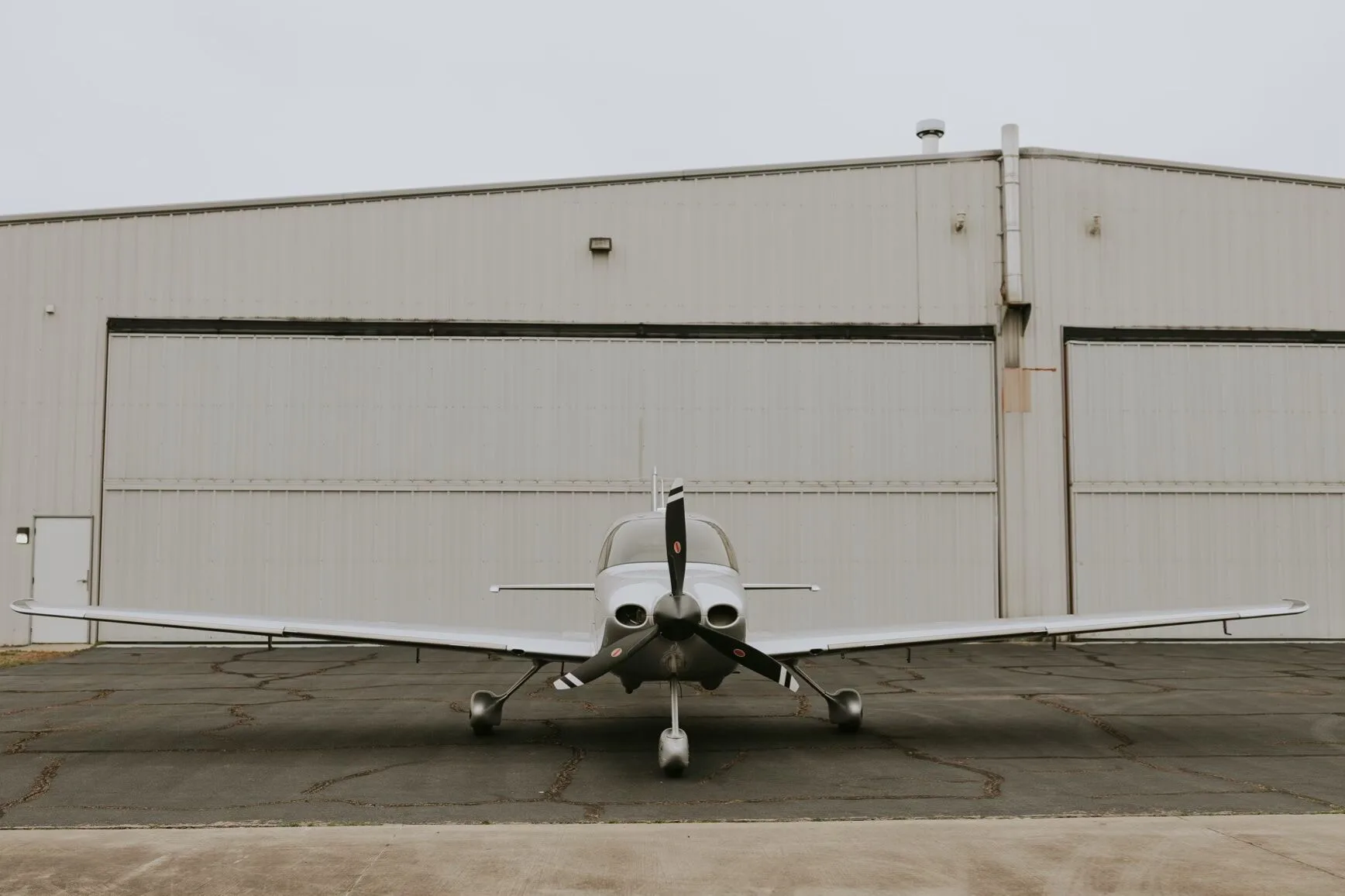 A head-on view of a silver Cirrus SR22 GTS aircraft with a black propeller, parked in front of a hangar.
