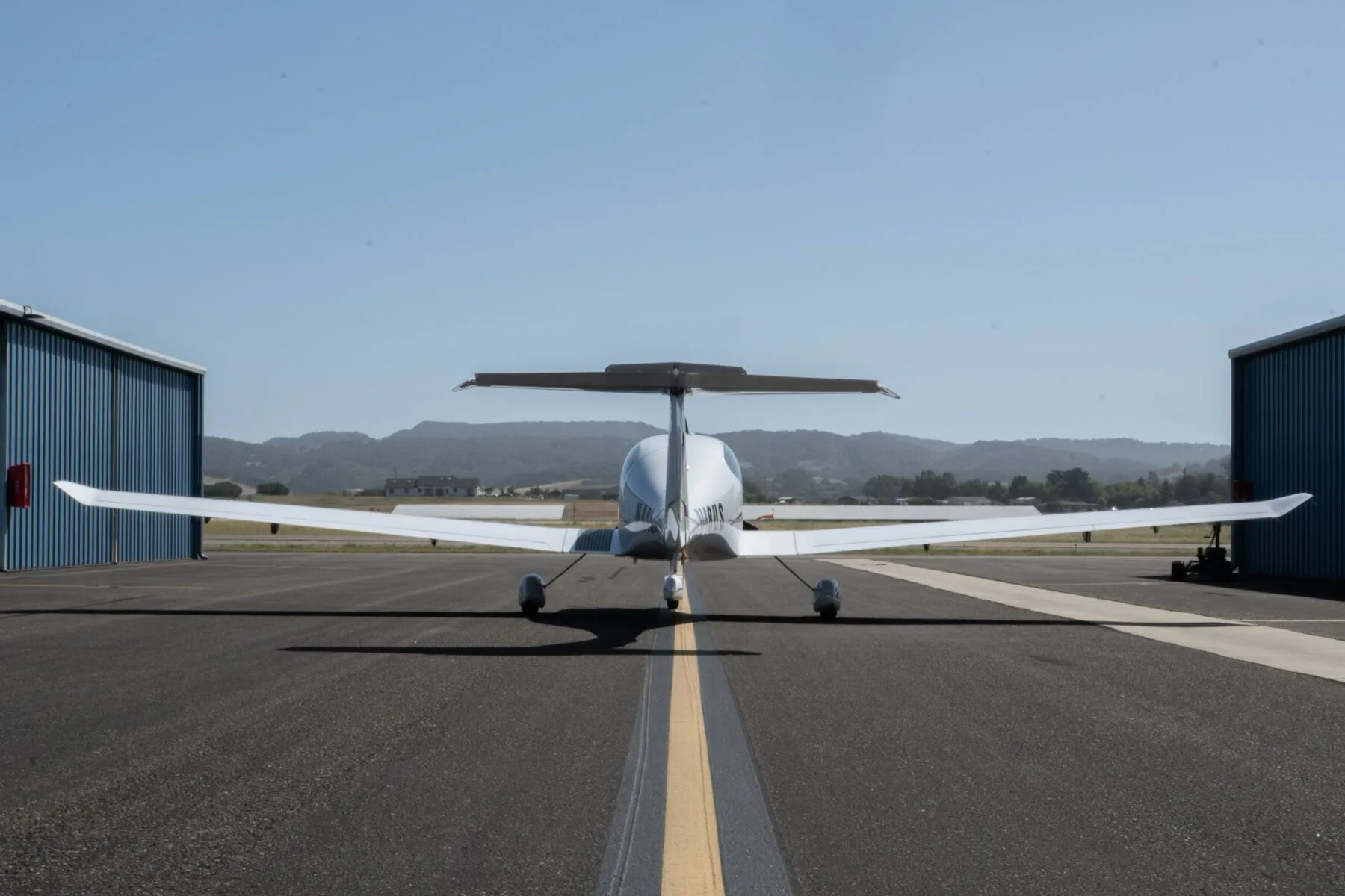 A white Diamond DA40 Star XL aircraft, registration N448US, viewed from the rear on an airport runway with hangars and hills in the background.
