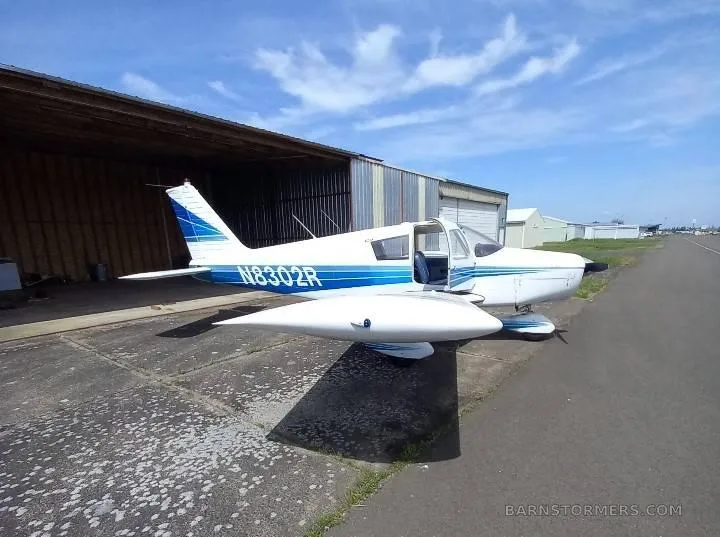 A white and blue Piper Cherokee 140 (N8302R) parked outside a hangar on a sunny day.