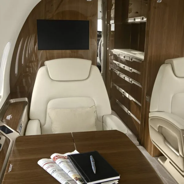 A close-up view of a private jet's cabin interior, showing a cream leather seat, a wooden table with a notebook, and storage cabinets.