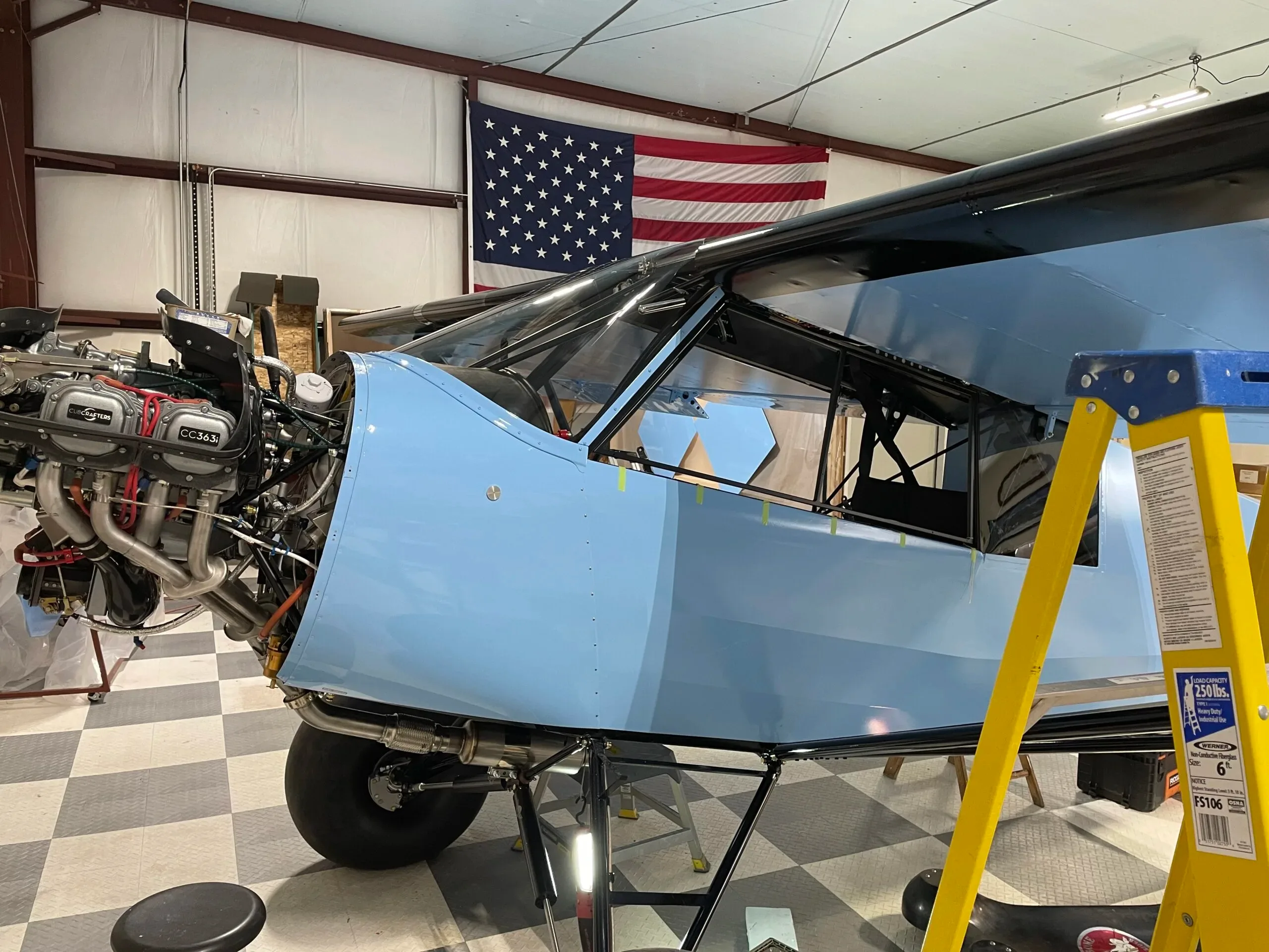 A light blue aircraft in a hangar with its engine cowling removed, revealing the engine, next to a yellow step ladder.