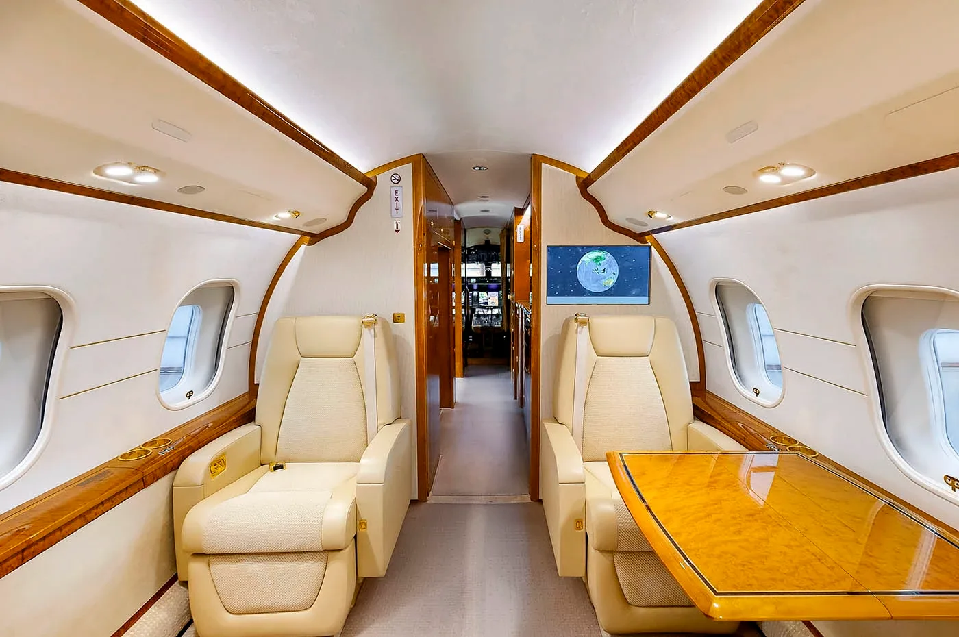 A section of the private jet's cabin with two beige reclining seats, a polished wood table, and a wall-mounted screen showing a globe.
