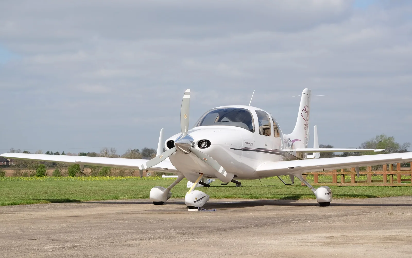 A white Cirrus SR22 aircraft with purple accents and a Snoopy graphic on the tail, parked on a tarmac with fields in the background.