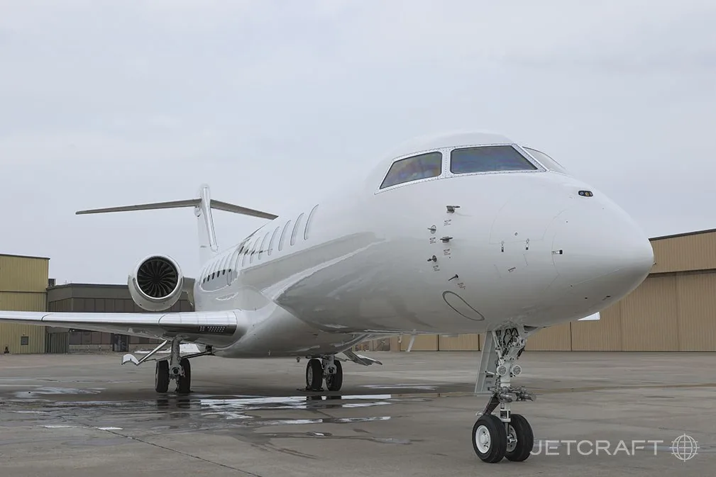 A white Bombardier Global 7500 business jet is parked on a wet tarmac, viewed from the front-right.