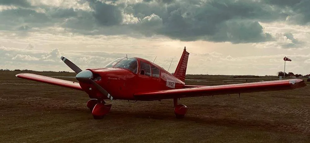 A red Piper Cherokee 180 (N8325W) with a propeller, parked on a grassy field under a cloudy sky.