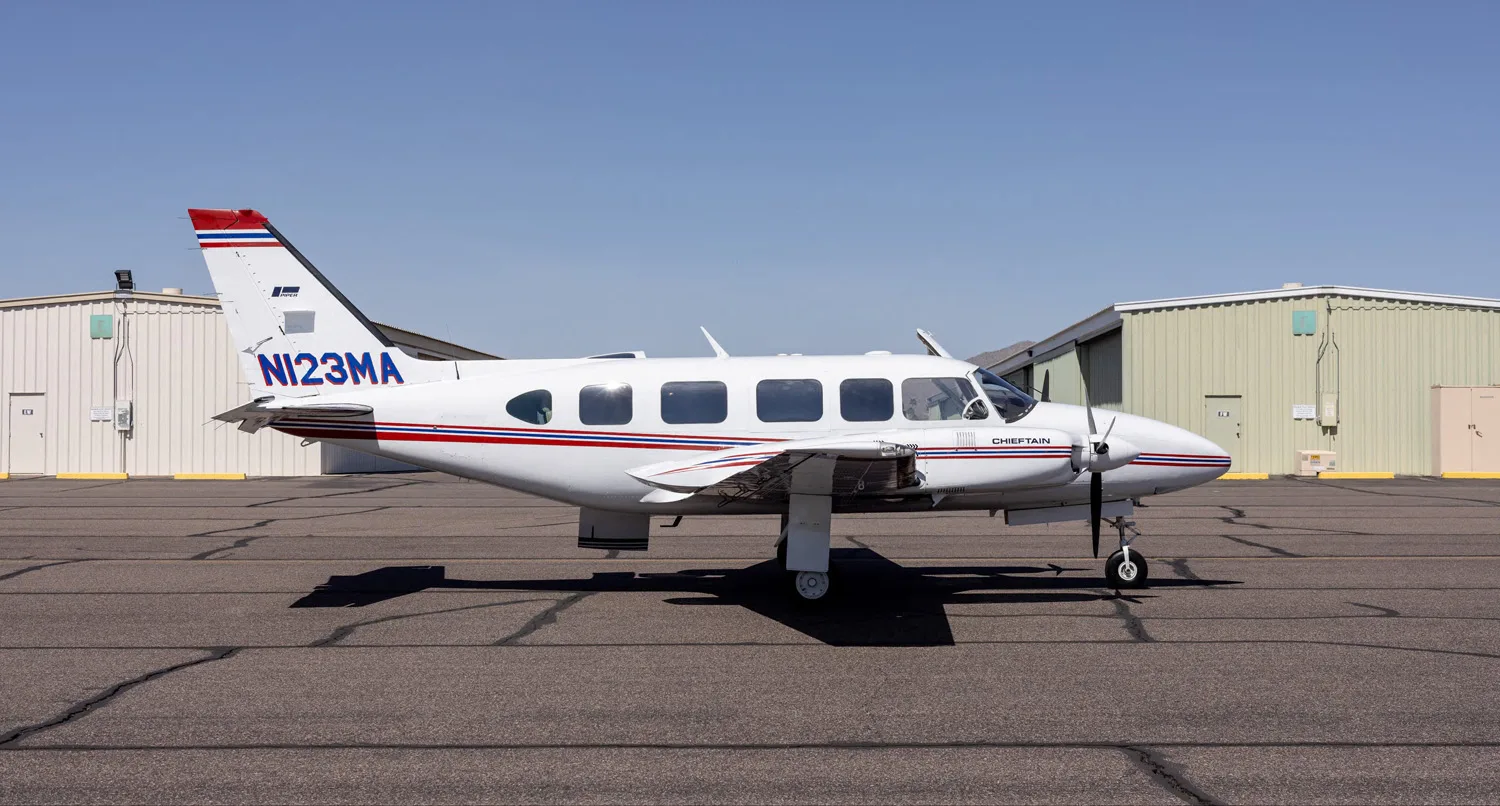 A white Piper Chieftain aircraft with red and blue stripes is parked on an asphalt tarmac with hangars in the background.