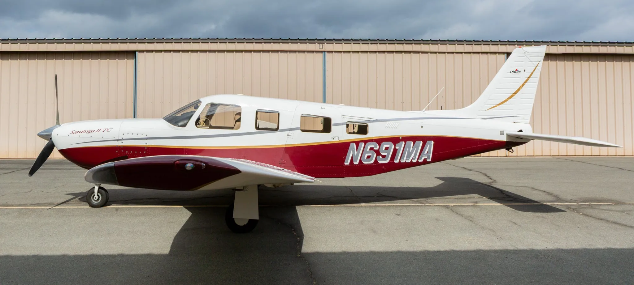 Side view of a white and maroon Piper Saratoga II TC aircraft, registration N691MA, parked in front of a hangar.