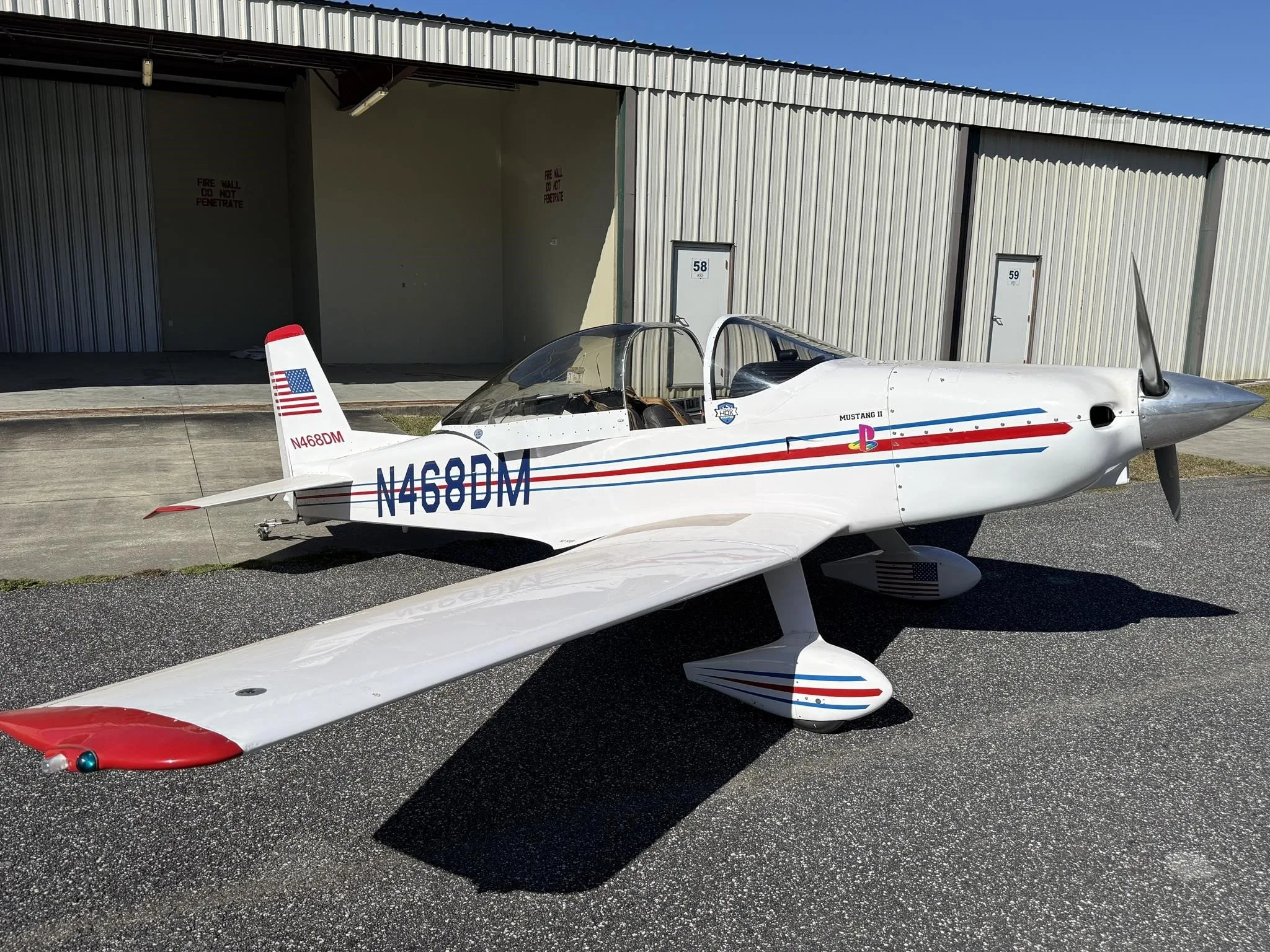 A white Mustang II aircraft with red wingtips and blue/red stripes, registration N468DM, parked in front of an open hangar.