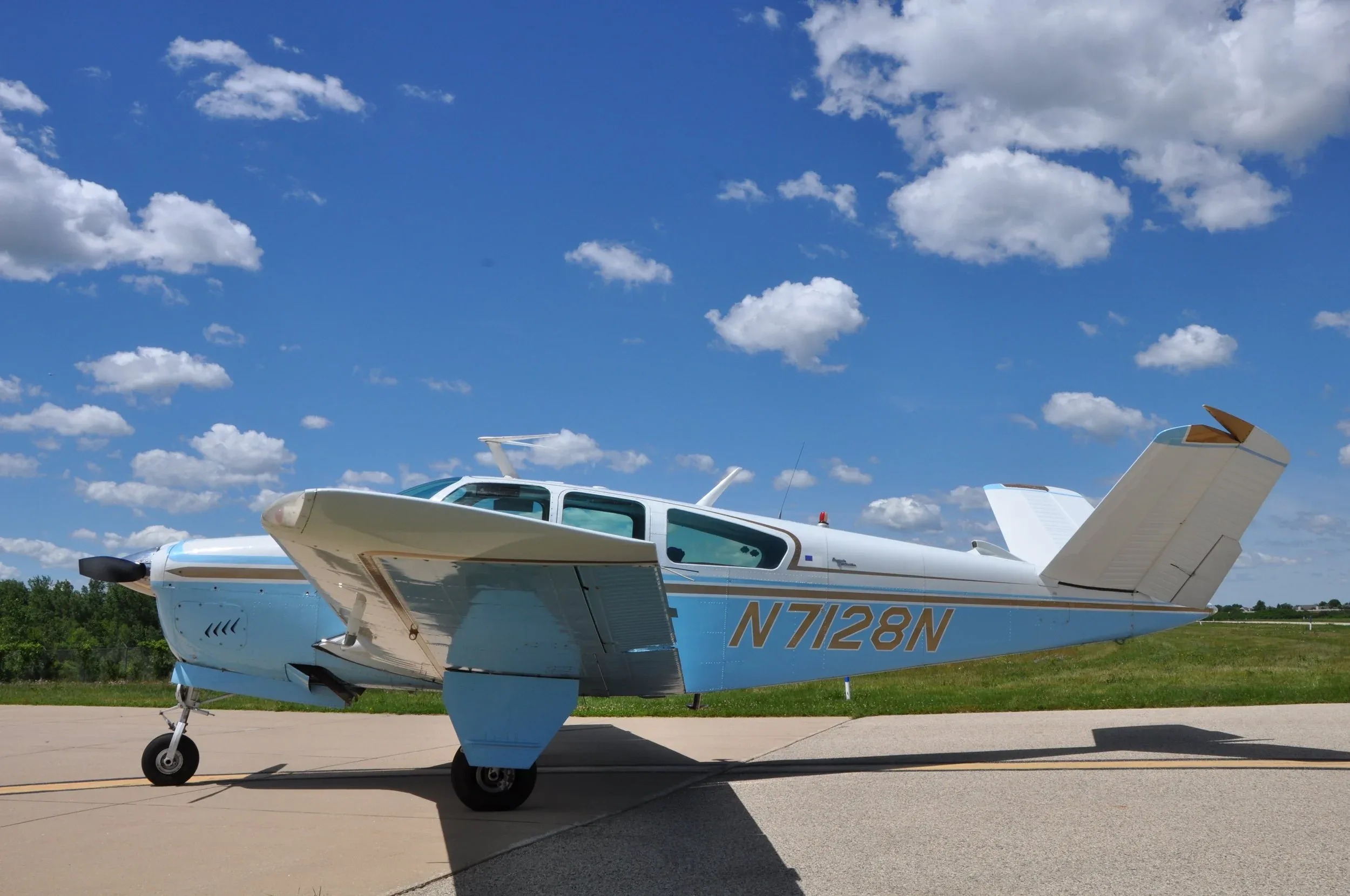 A side view of a light blue and white single-engine aircraft with a V-tail, showing the registration N7128N, parked on an asphalt runway.