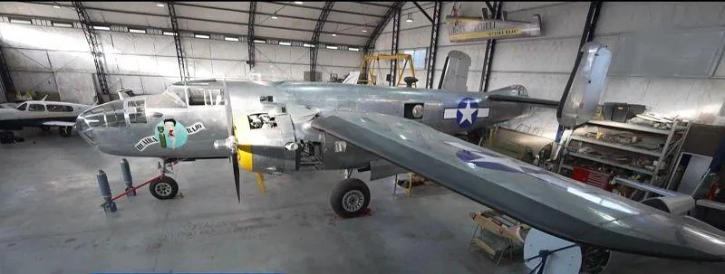 A full side view of a silver B-25 Mitchell bomber with US star insignia on the fuselage and wing, parked in a hangar.