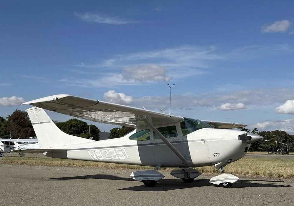 A white Cessna 172 with gray stripes, registration N92351, parked on an airfield under a blue sky with clouds.