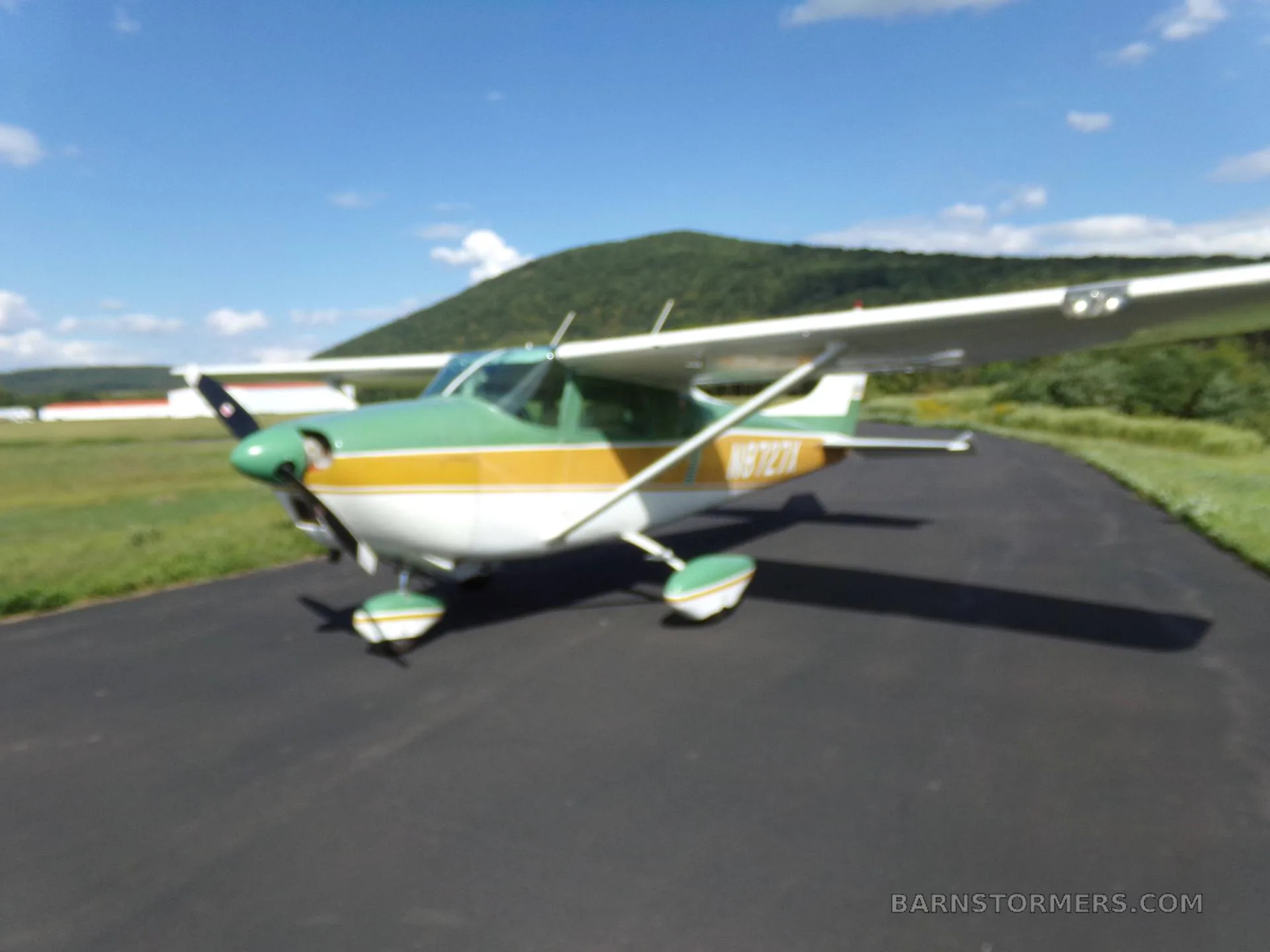 A green, gold, and white Cessna 172 aircraft with registration N8727X, viewed from the front-right on an asphalt ramp under a clear blue sky.