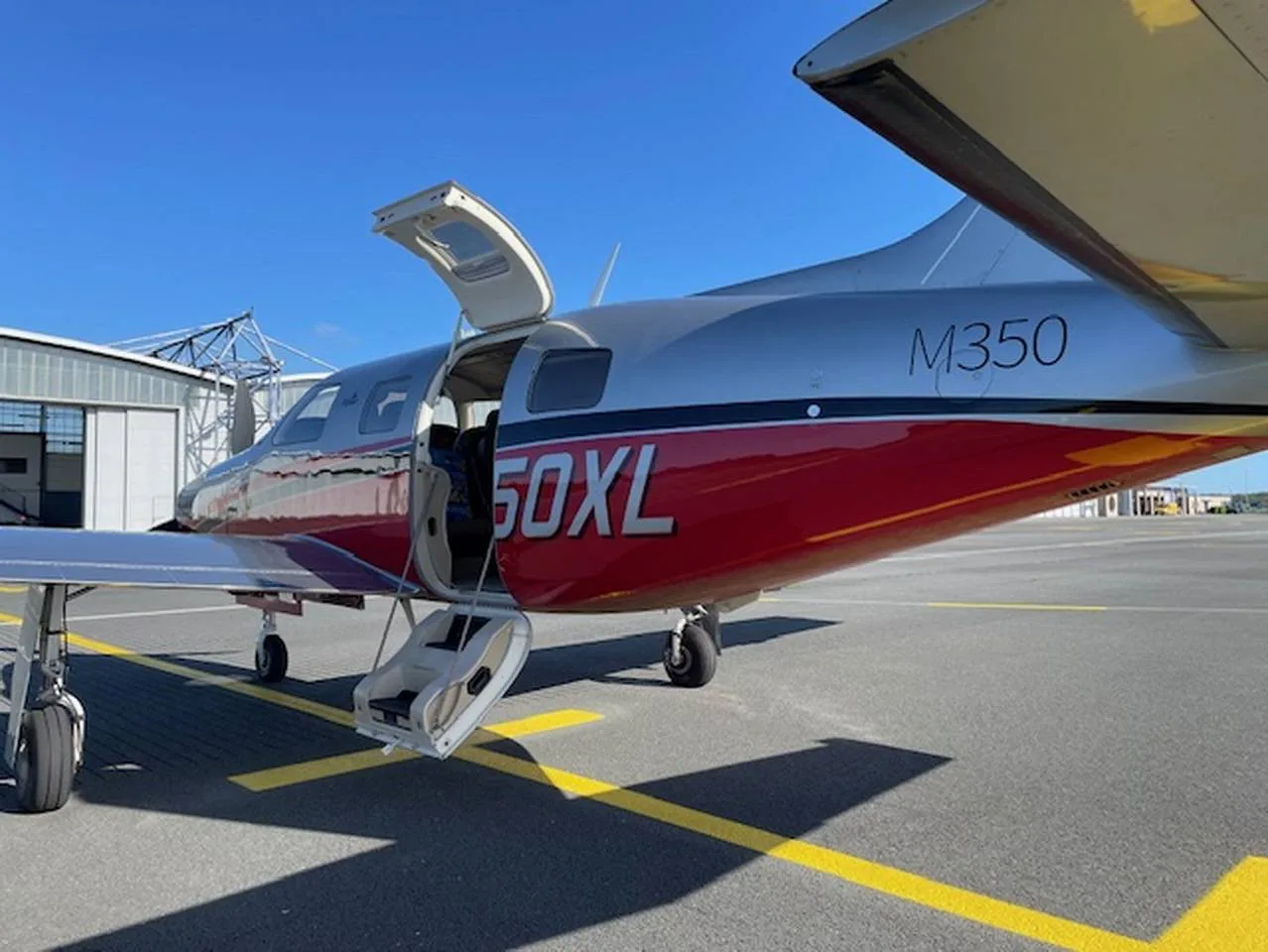 A silver and red Piper M350 aircraft with its main door open, parked on an airport tarmac under a clear blue sky.