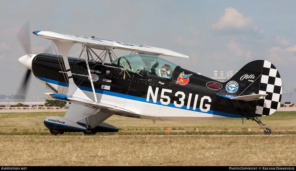 A black, white, and blue Pitts biplane with registration N5311G is taxiing on a grassy airfield with a pilot waving.