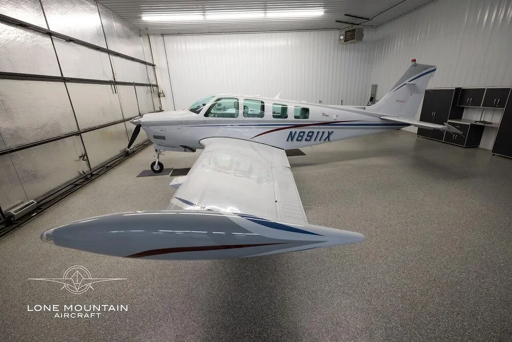 A white Beechcraft Bonanza A36 aircraft with blue and red stripes, viewed from the rear right in a hangar.