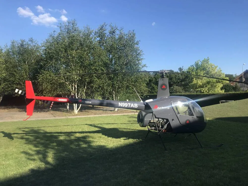 A dark gray Robinson R44 helicopter, N997AB, with red tail accents, parked on a grassy area with trees in the background.