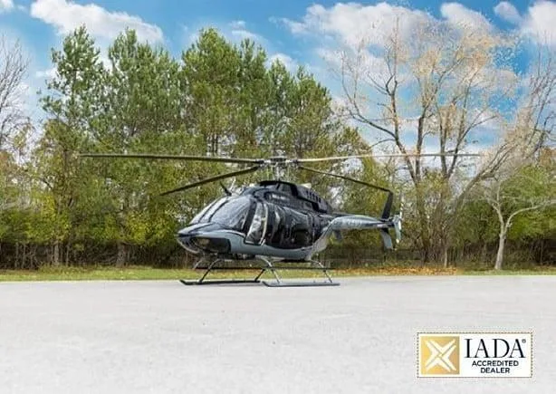 A black and grey Bell 407 helicopter is parked on a concrete pad with trees in the background under a blue sky.