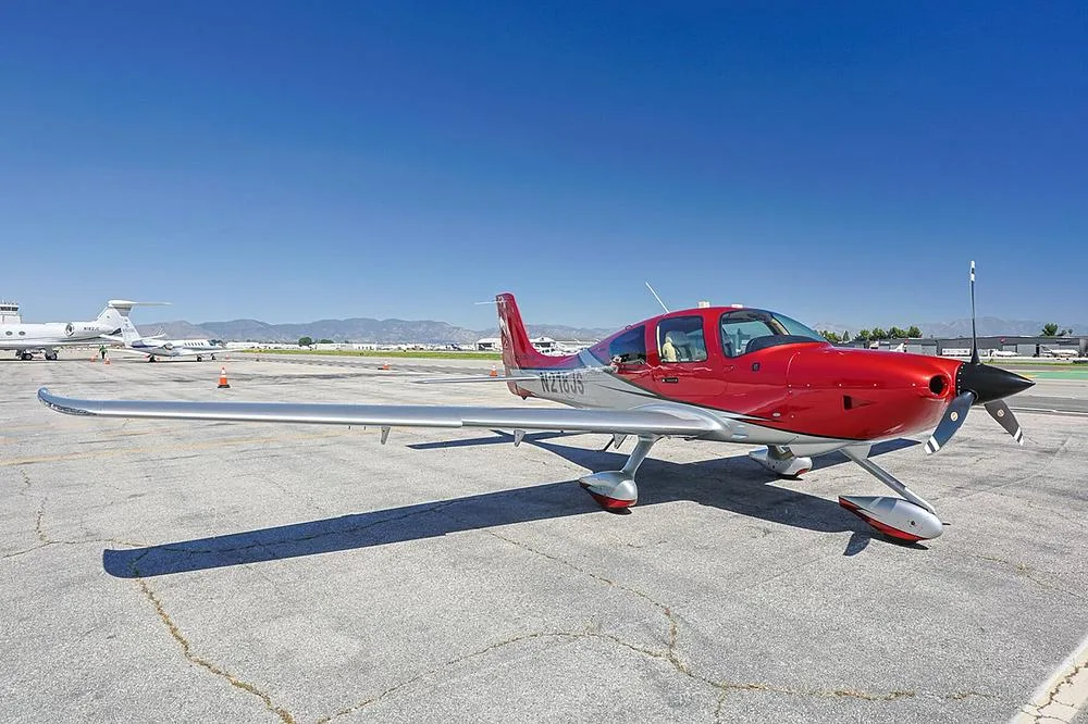 A red and silver Cirrus SR22 aircraft with registration N218JS parked on an airport tarmac under a clear blue sky.