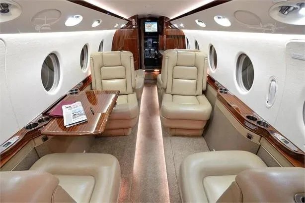 The private jet's cabin interior, featuring beige leather seats, polished wood accents, and a view towards the cockpit.
