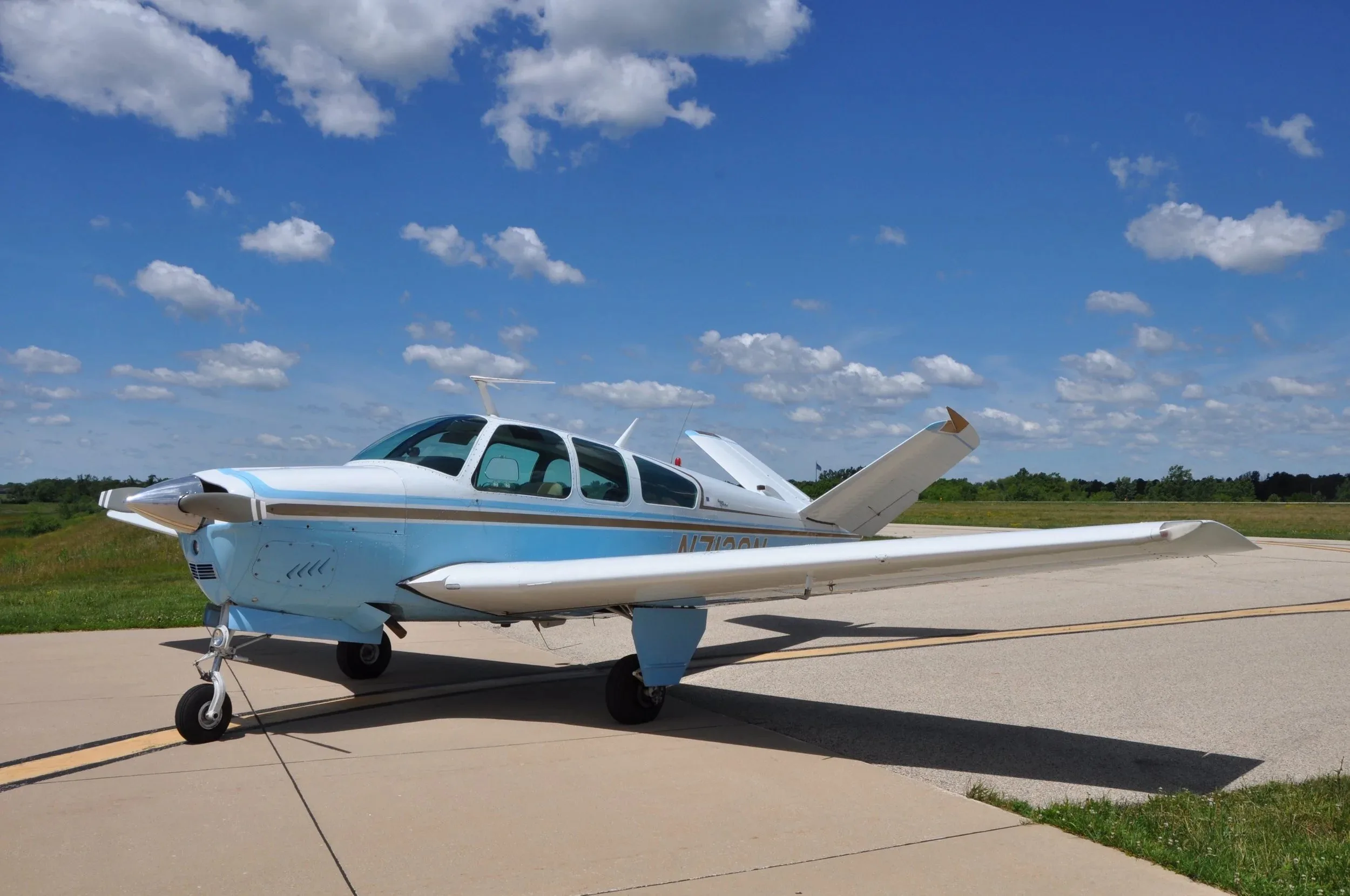 A light blue and white Beechcraft Bonanza N7128N parked on an airfield under a blue sky with clouds.