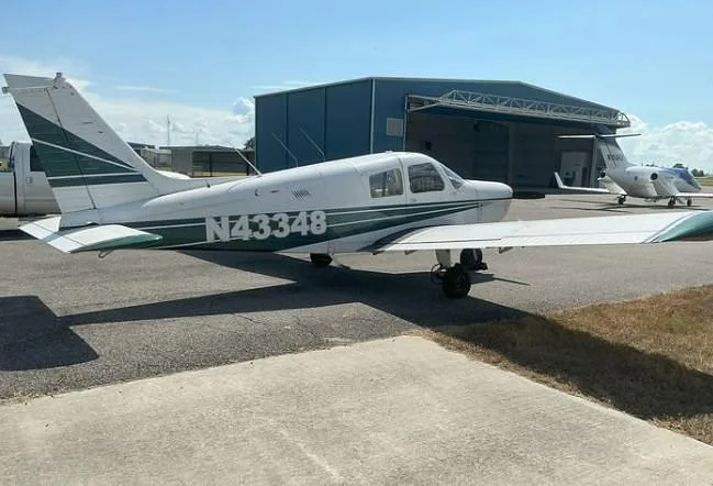 A white Piper Cherokee with green stripes, registration N43348, parked on an airfield with a hangar and another jet in the background.
