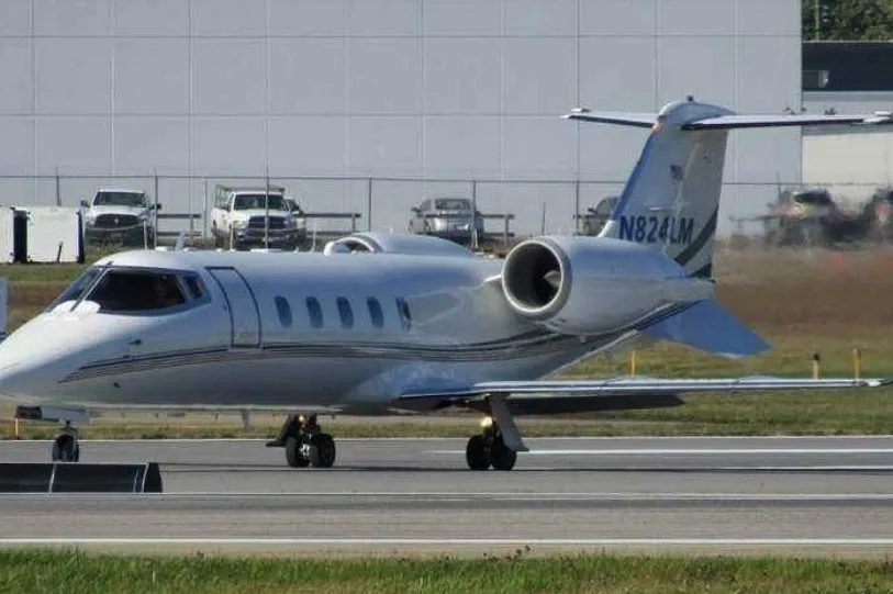 A white business jet with blue and gold stripes, registration N824LM, taxiing on a runway with industrial buildings in the background.
