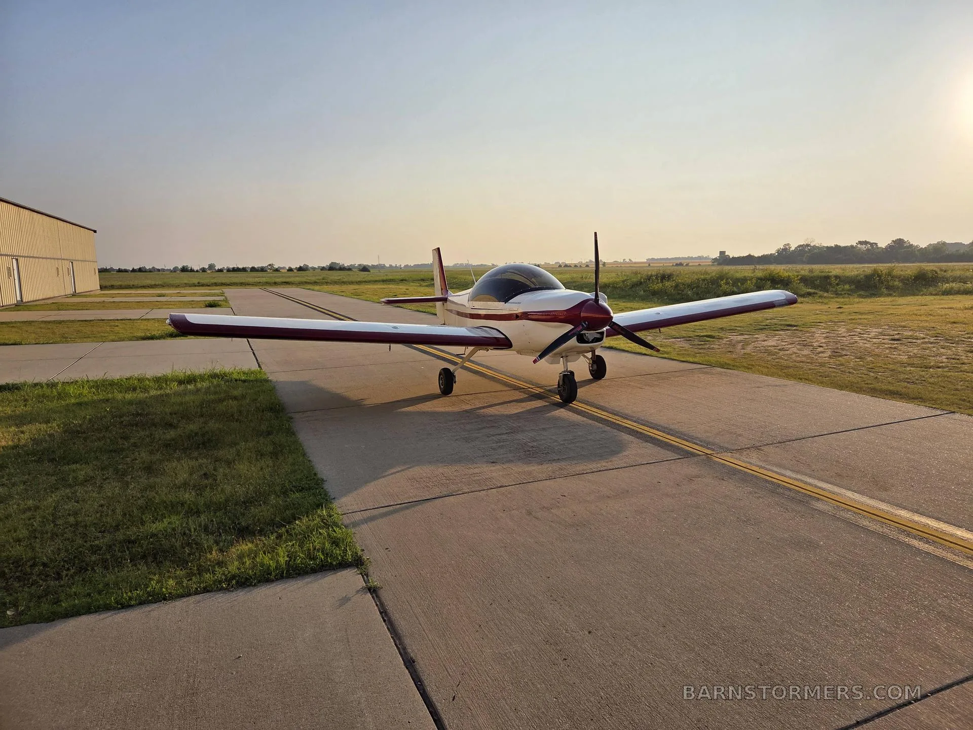 A white and red single-engine aircraft with a bubble canopy is parked on an asphalt taxiway at sunset.