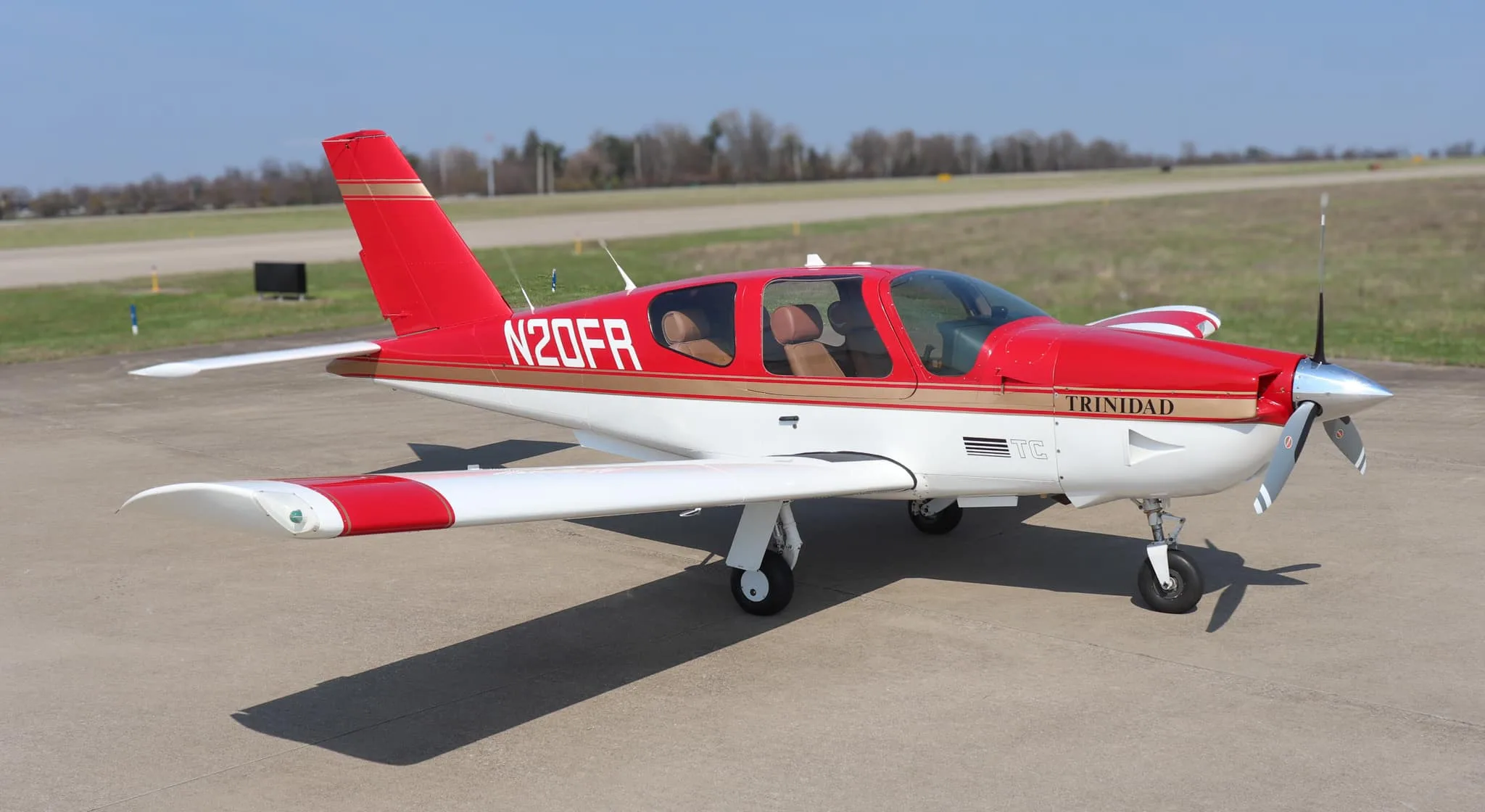 A red and white Socata Trinidad aircraft with gold stripes is parked on an airport tarmac, viewed from the front-right.