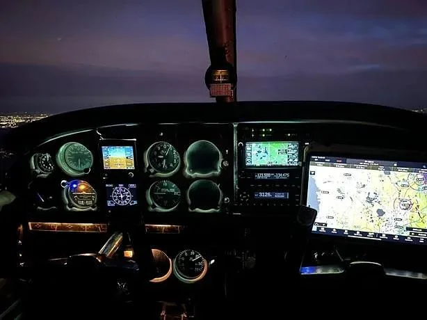 An illuminated aircraft cockpit at night, showing the instrument panel with various gauges and digital displays, and a view of city lights outside.