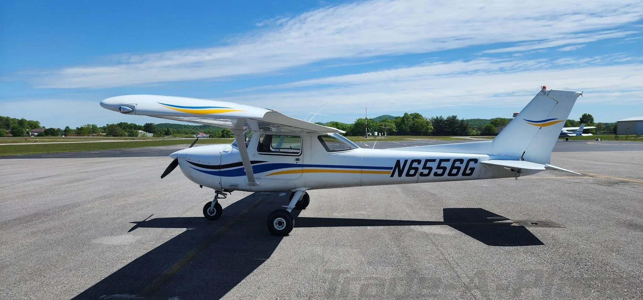 A white Cessna 150 with blue and yellow stripes, parked on an airport tarmac under a partly cloudy sky.