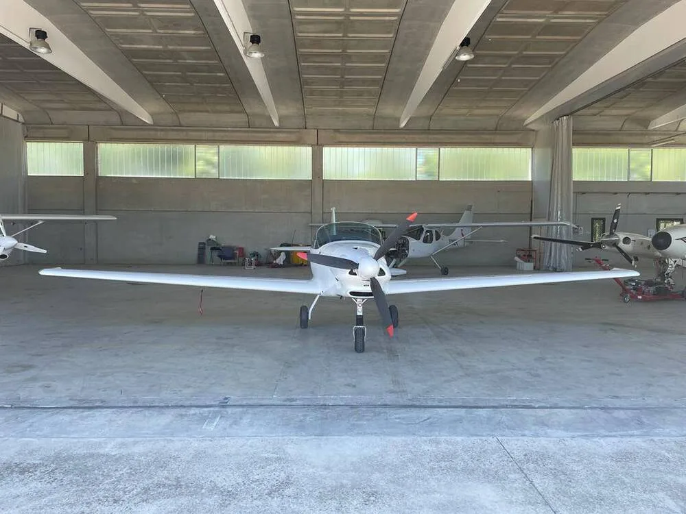 A white Fascination ultralight aircraft with a clear canopy is parked inside a hangar, viewed head-on.