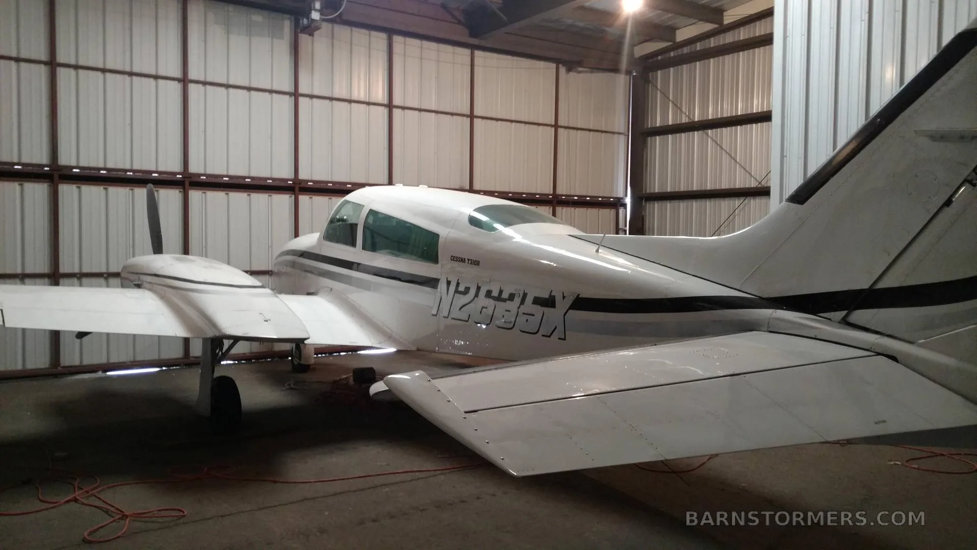 The rear-left view of a white Cessna 310 twin-engine aircraft with black stripes parked inside a metal hangar.
