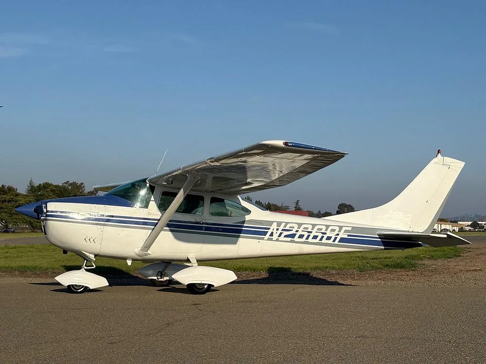 A white and blue Cessna 182 aircraft (N2668F) parked on an airfield with trees and buildings in the background.