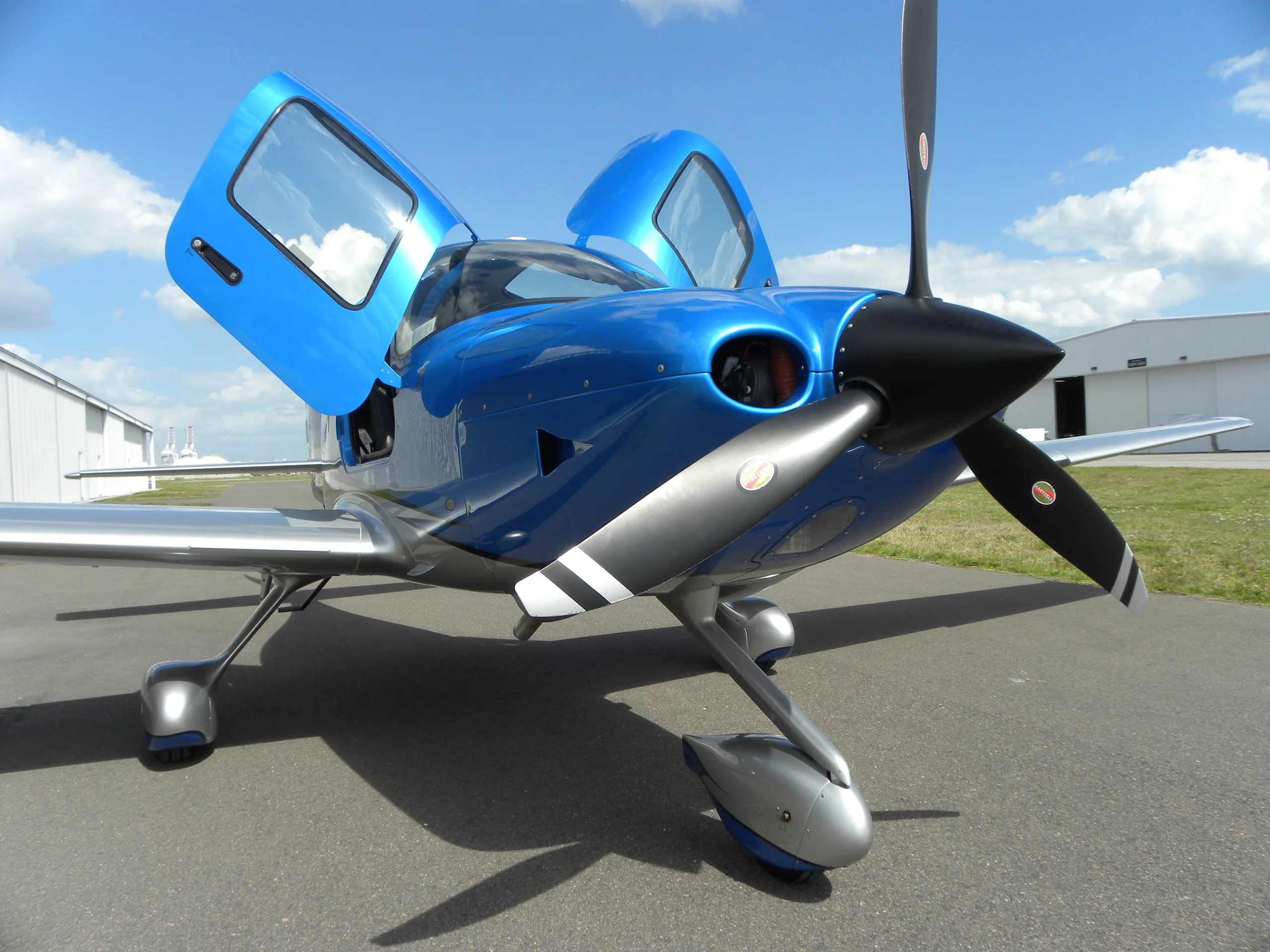 A blue and silver single-engine propeller aircraft with open gull-wing doors is parked on an airfield.