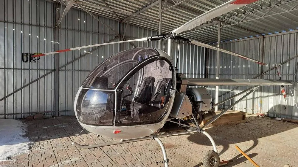 A silver AK1-3 helicopter is parked inside a metal hangar, viewed from the front-left.