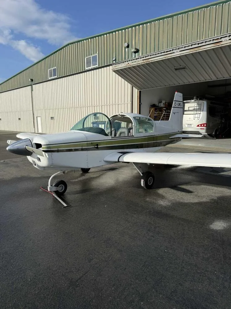 A white and green Grumman American AA-5 Traveler aircraft with registration C-GWES parked outside a hangar with its canopy open.