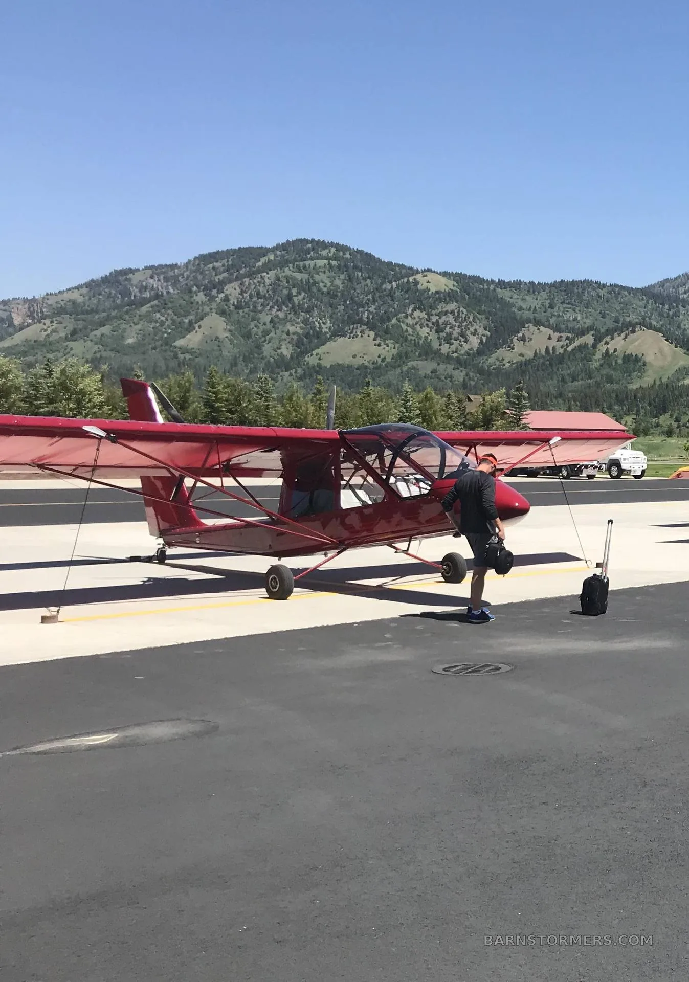 A red Air Cam aircraft is parked on an airfield with a person and luggage, set against a backdrop of green mountains.