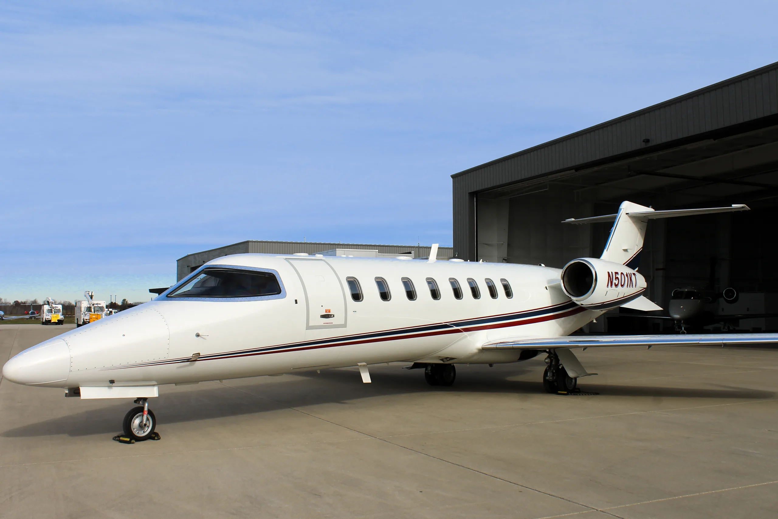 A white Learjet business jet with red and black stripes parked on a tarmac next to a hangar.