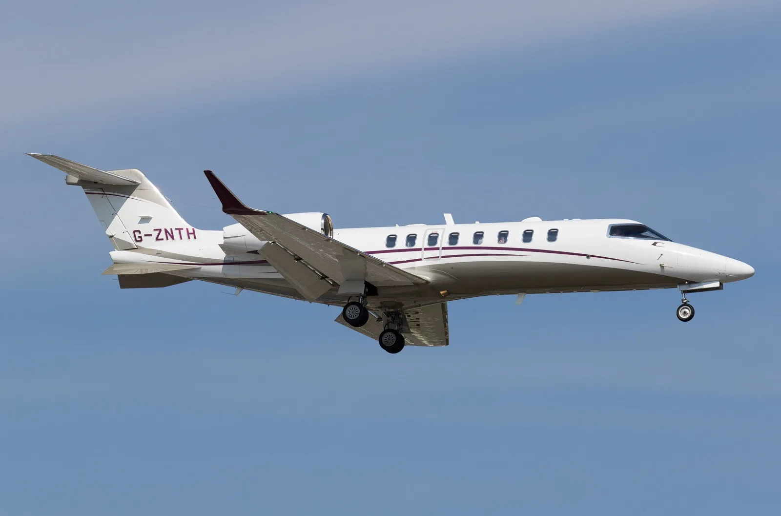 A white business jet with maroon accents and registration G-ZNTH is shown in flight with landing gear extended against a blue sky.