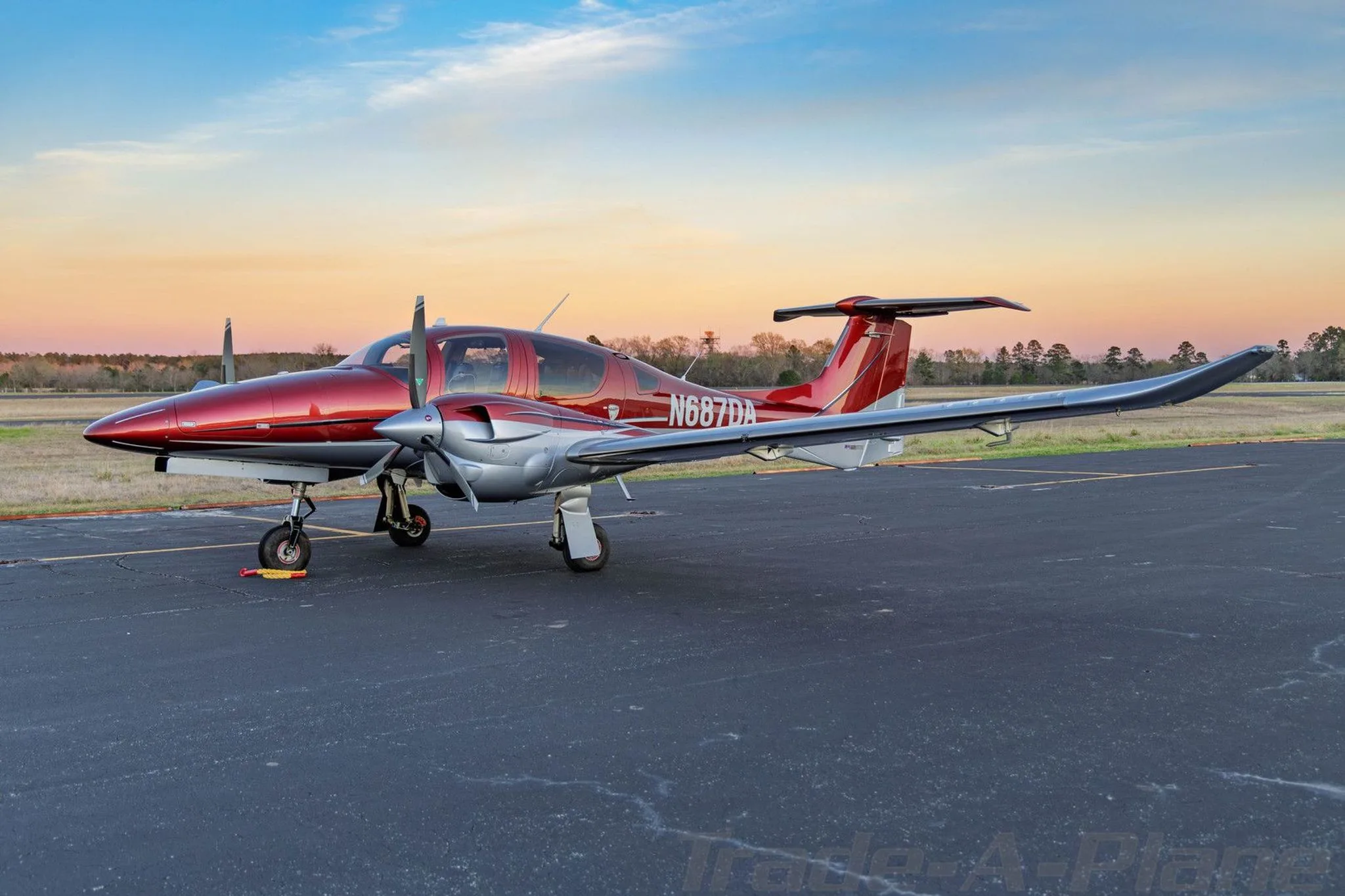 A side view of a red and silver Diamond DA62 aircraft parked on an airfield at sunset.